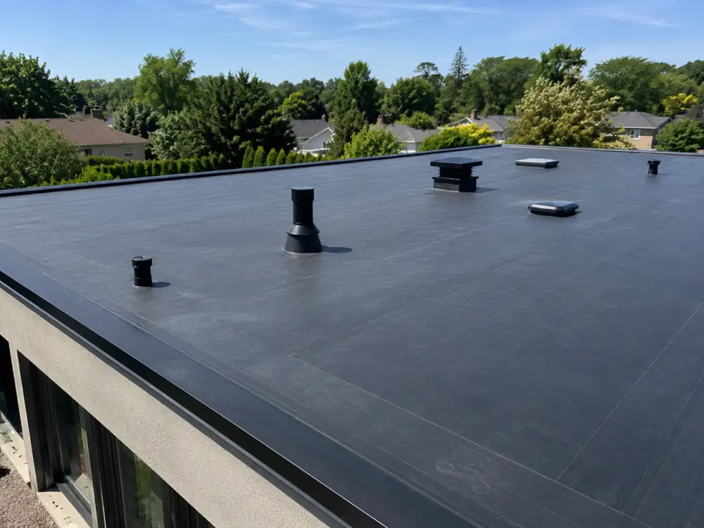 Black EPDM rubber membrane on a flat roof with vent stacks and hatches, treed yards in the background