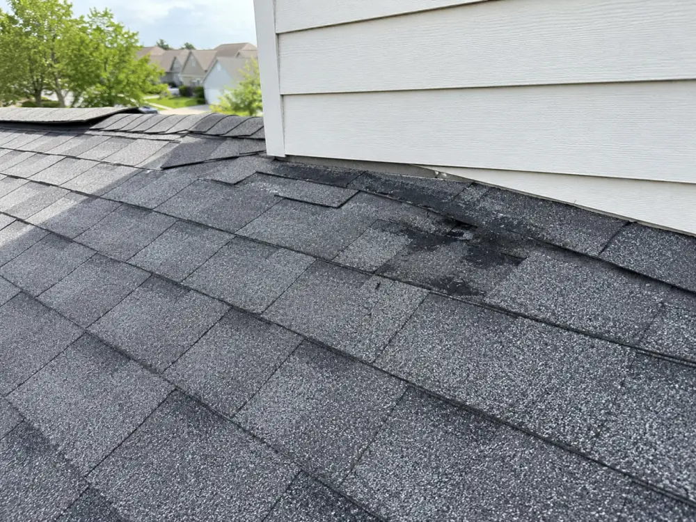 Close-up of a grey asphalt shingle roof against white siding, with darker mismatched shingles and worn patches at the sidewall