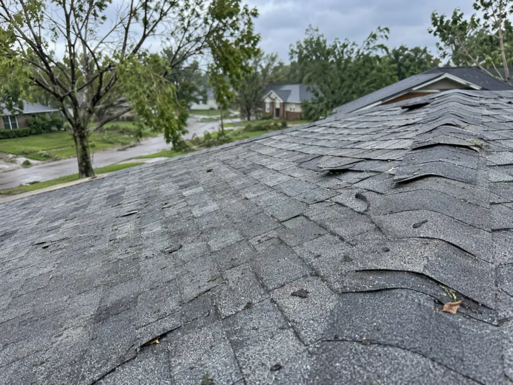 Asphalt shingle roof with shingles lifted and creased along the hip after a storm, wet street and downed branches below